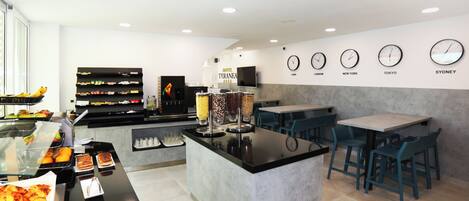 Un espace pour le petit-déjeuner à l’hôtel, avec un buffet, des tables et des chaises, ainsi que des horloges indiquant l’heure de différentes villes.