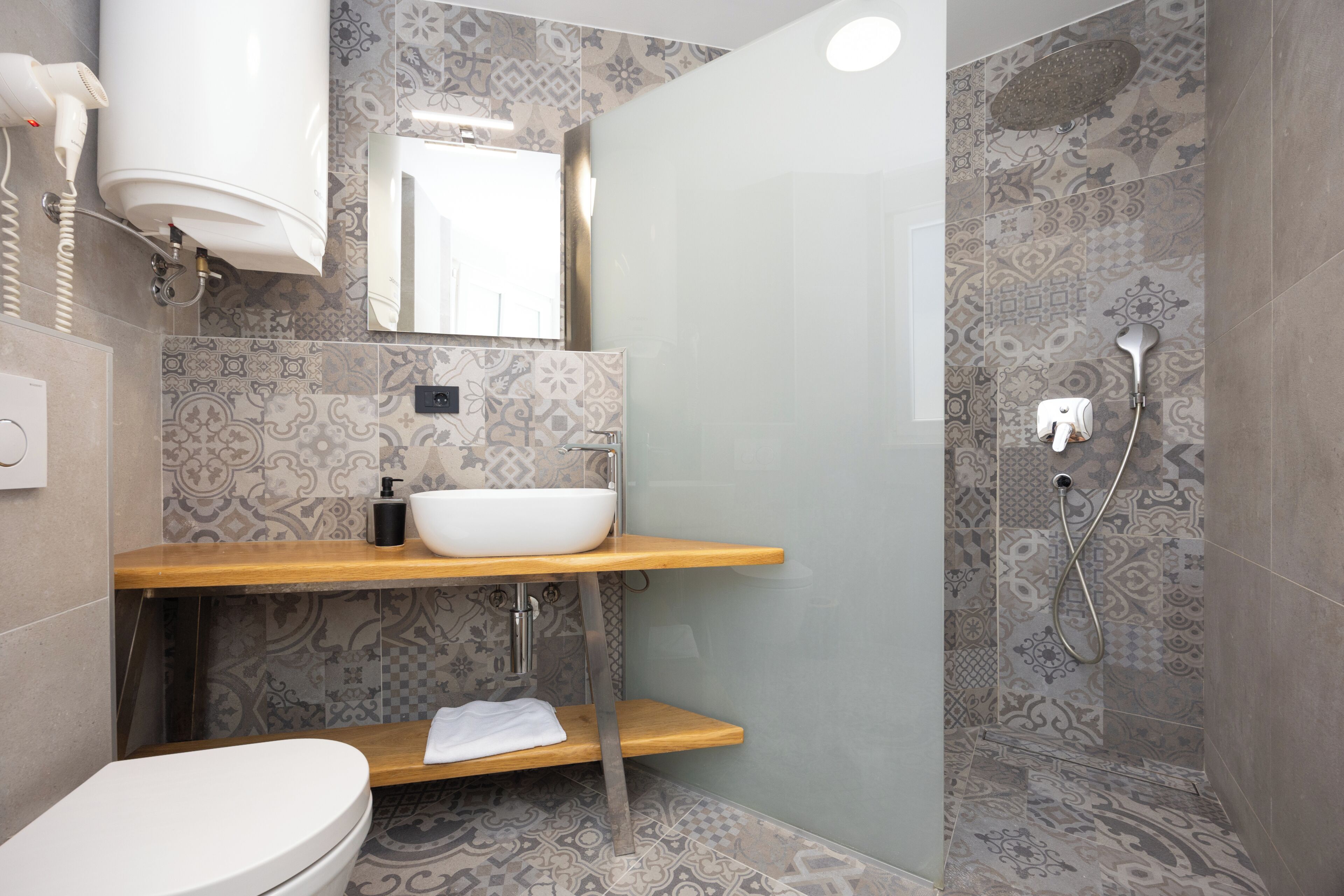 A modern bathroom with a white toilet, a wooden vanity with a white sink, a glass shower enclosure, and patterned tiles.