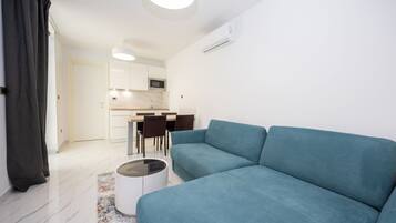 Una sala de estar moderna con un sofá color turquesa, una mesa de comedor con sillas, una zona de cocina y un equipo de aire acondicionado blanco.