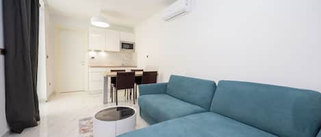 Una sala de estar moderna con un sofá color turquesa, una mesa de comedor con sillas, una zona de cocina y un equipo de aire acondicionado blanco.