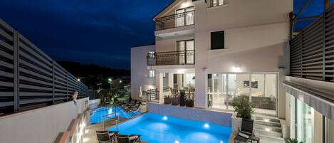 A modern two-story building with a swimming pool, outdoor seating, and a well-lit balcony.