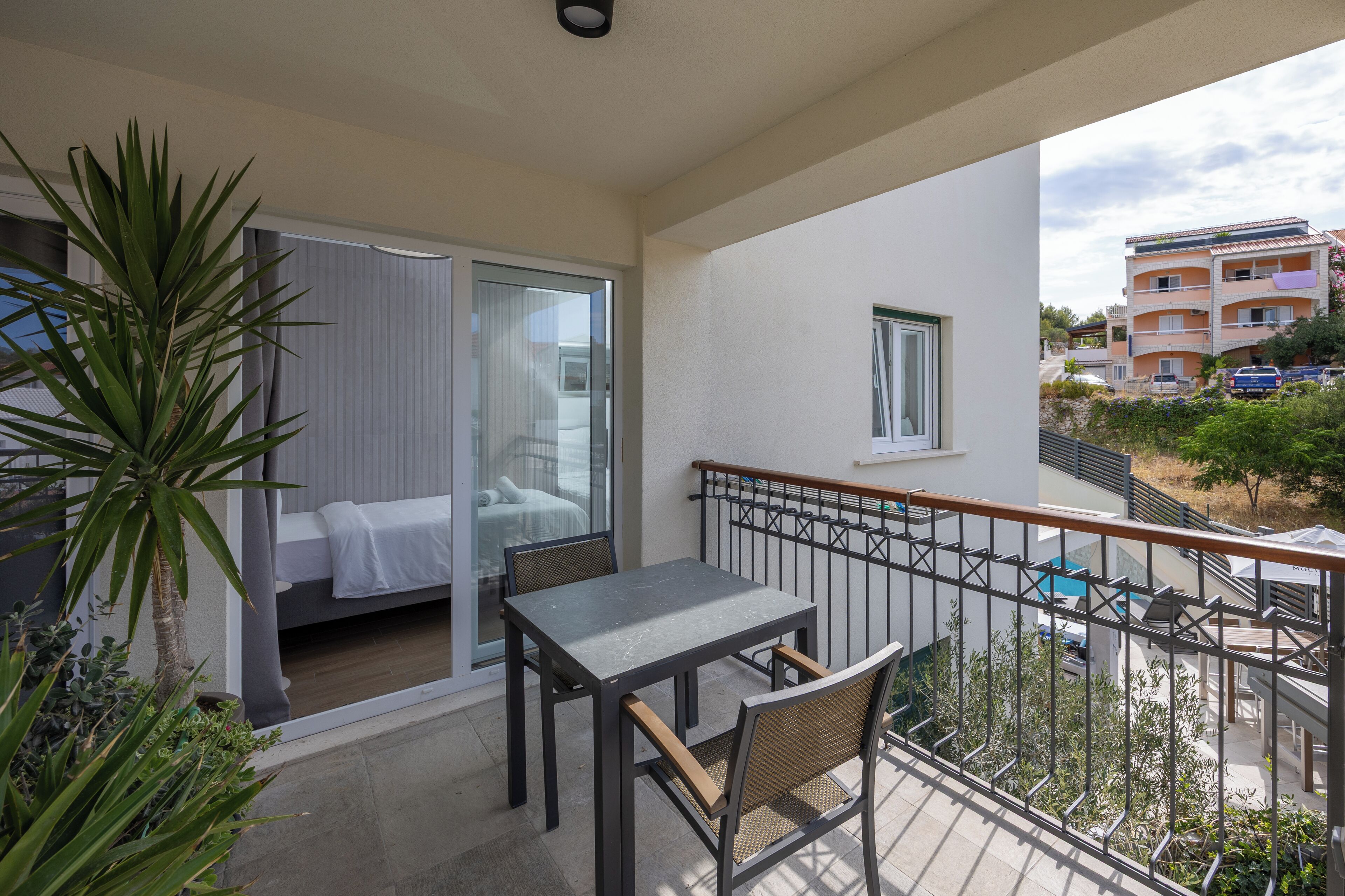 A balcony with a table and chairs, a bed with a white cover, and a view of a residential area with buildings and a pool.
