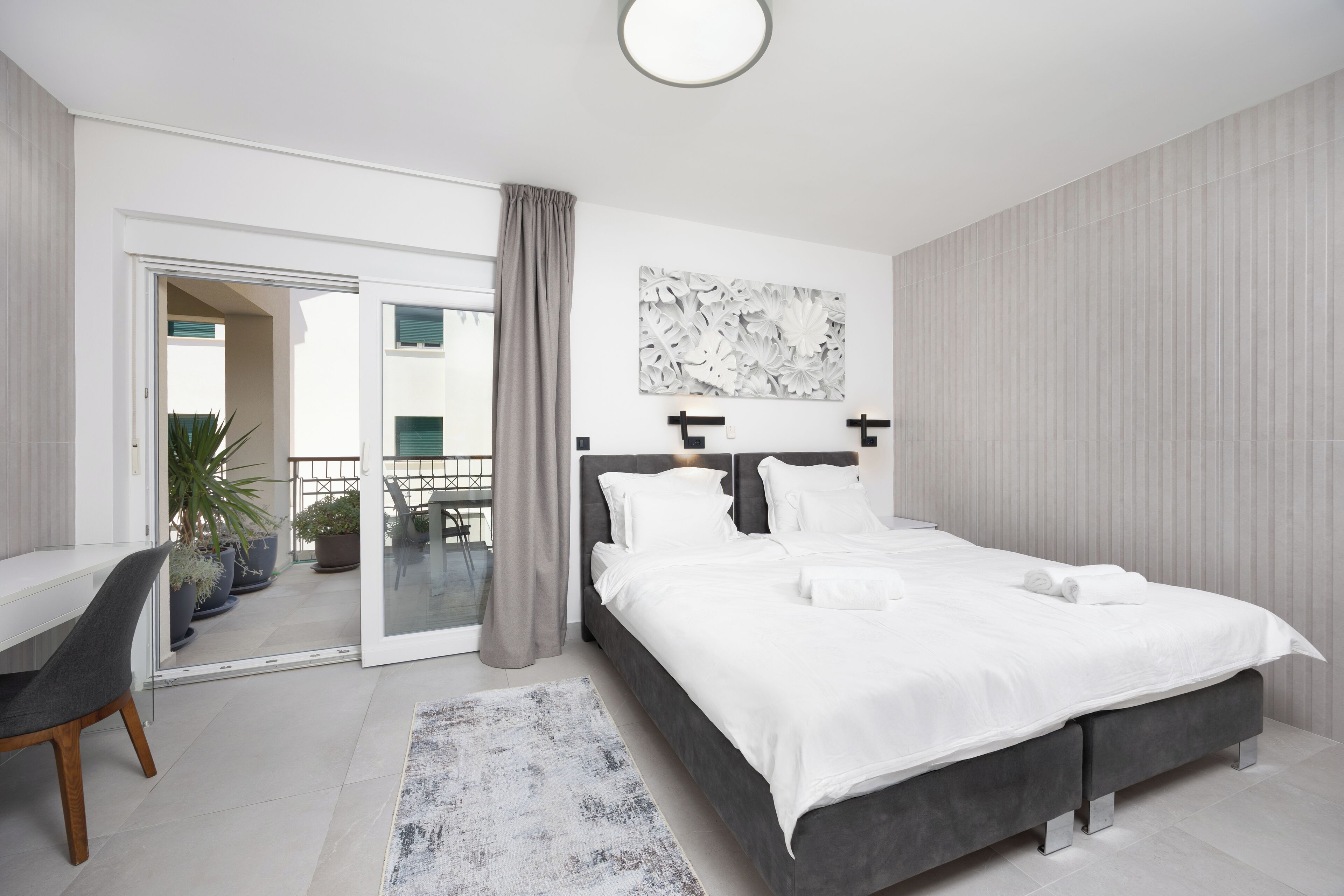A modern bedroom with a large bed, a desk, and a sliding glass door leading to a balcony.