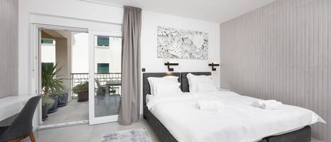 A modern bedroom with a large bed, a desk, and a sliding glass door leading to a balcony.