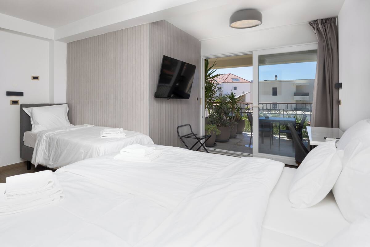 Habitación de hotel con dos camas, televisión, balcón con plantas en macetas y un ordenador portátil sobre una mesita.