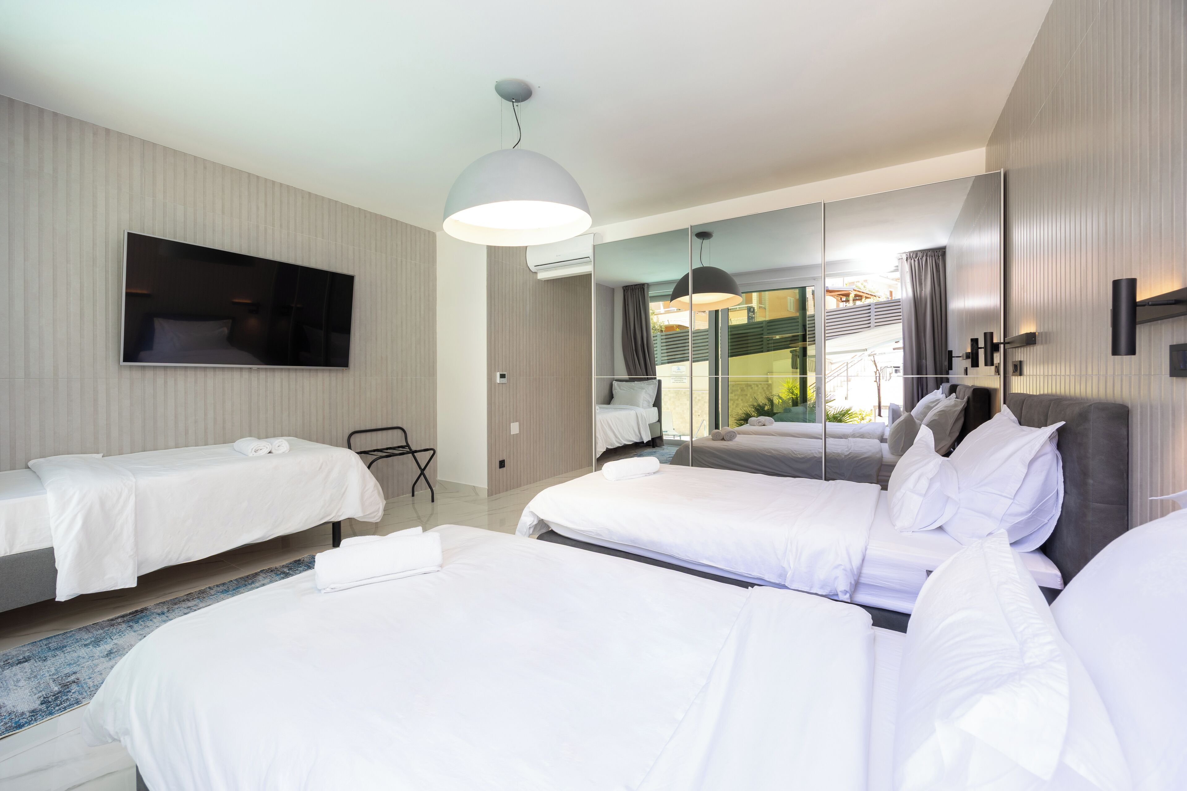 A modern hotel room with two single beds, a large mirror, a flat-screen TV, and a large window offering a view of the outdoors.