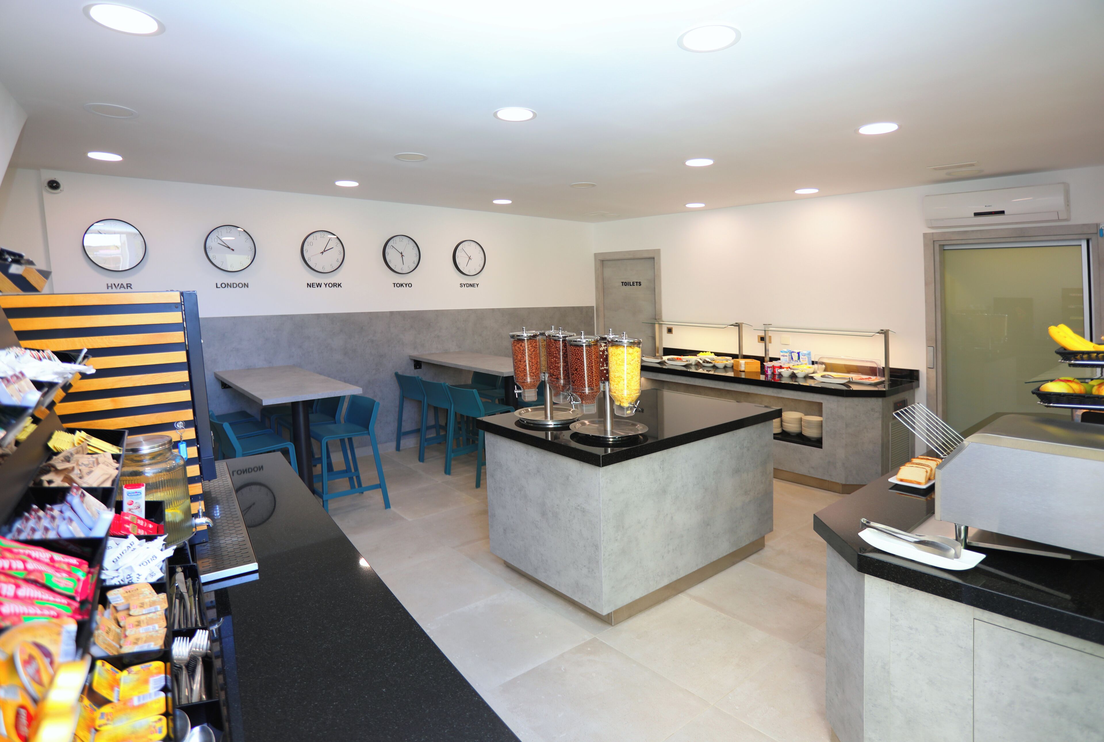 Un café moderne avec un comptoir, un espace salon et un mur orné d’horloges indiquant l’heure de différentes villes.