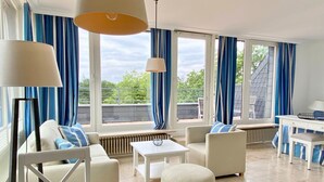 A modern living room with a sofa, ottoman, and coffee table, large windows with blue curtains, and a view of the outdoors.