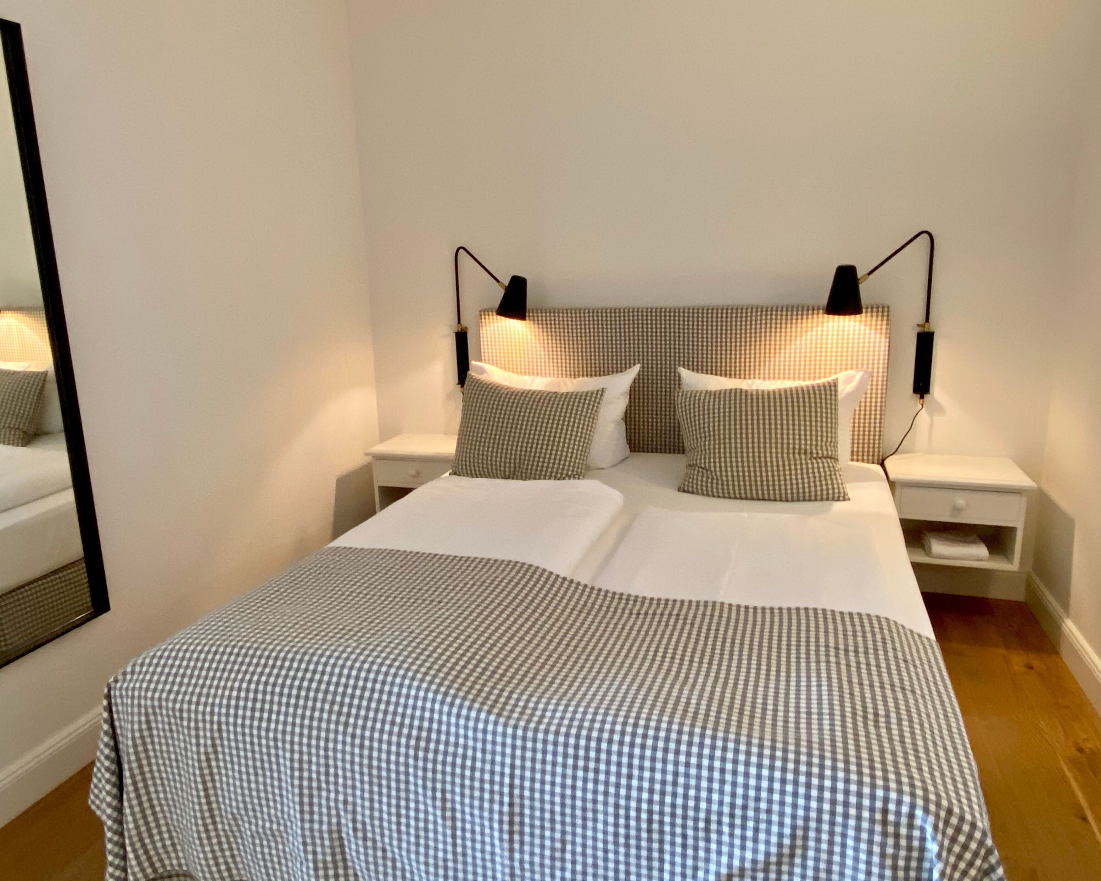 A neatly made bed with a checkered bedspread and pillows, two bedside tables with lamps, and a mirror on the left wall.