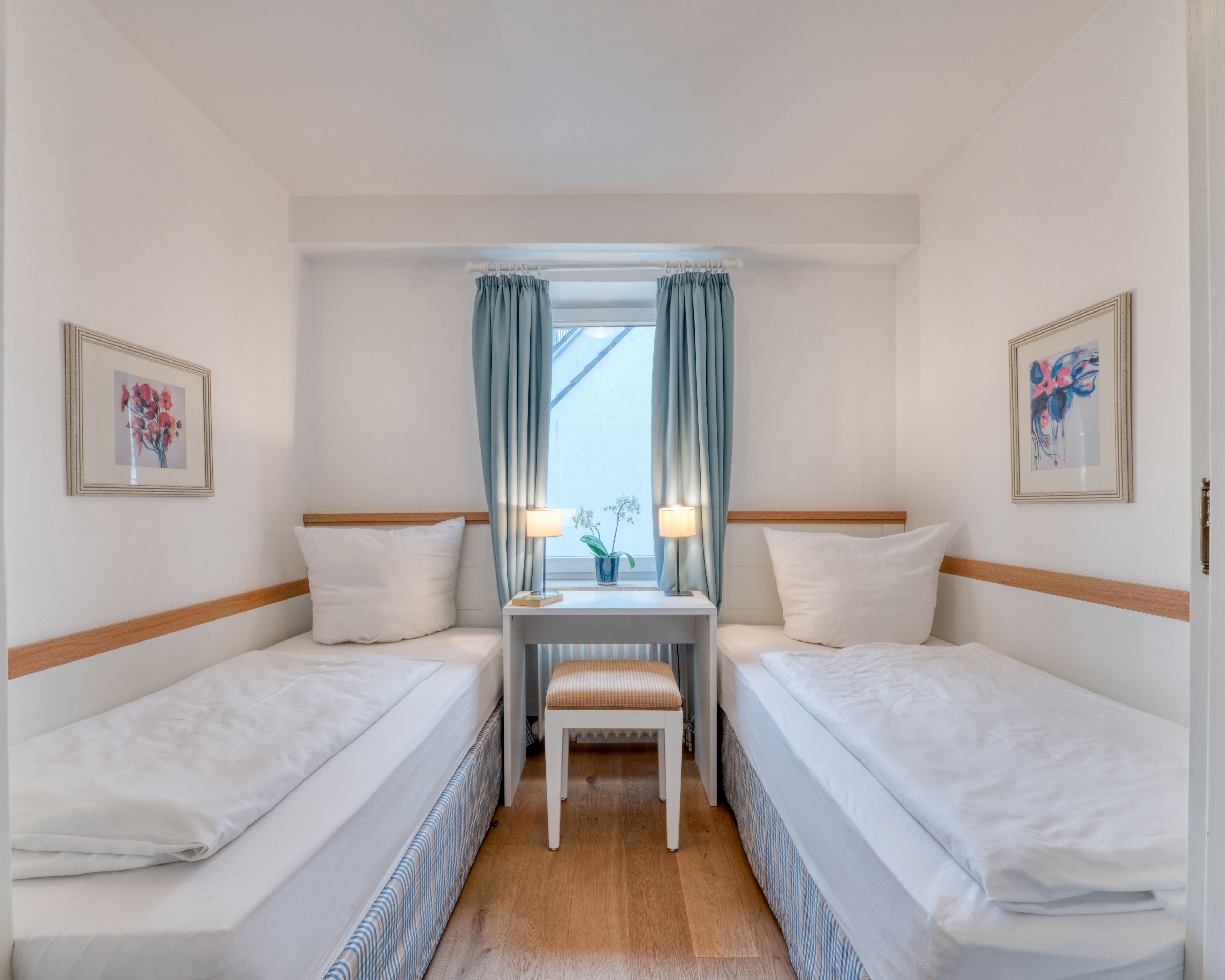 Camera d'albergo con due letti singoli, un tavolino con una lampada, una sedia e una finestra con tende.