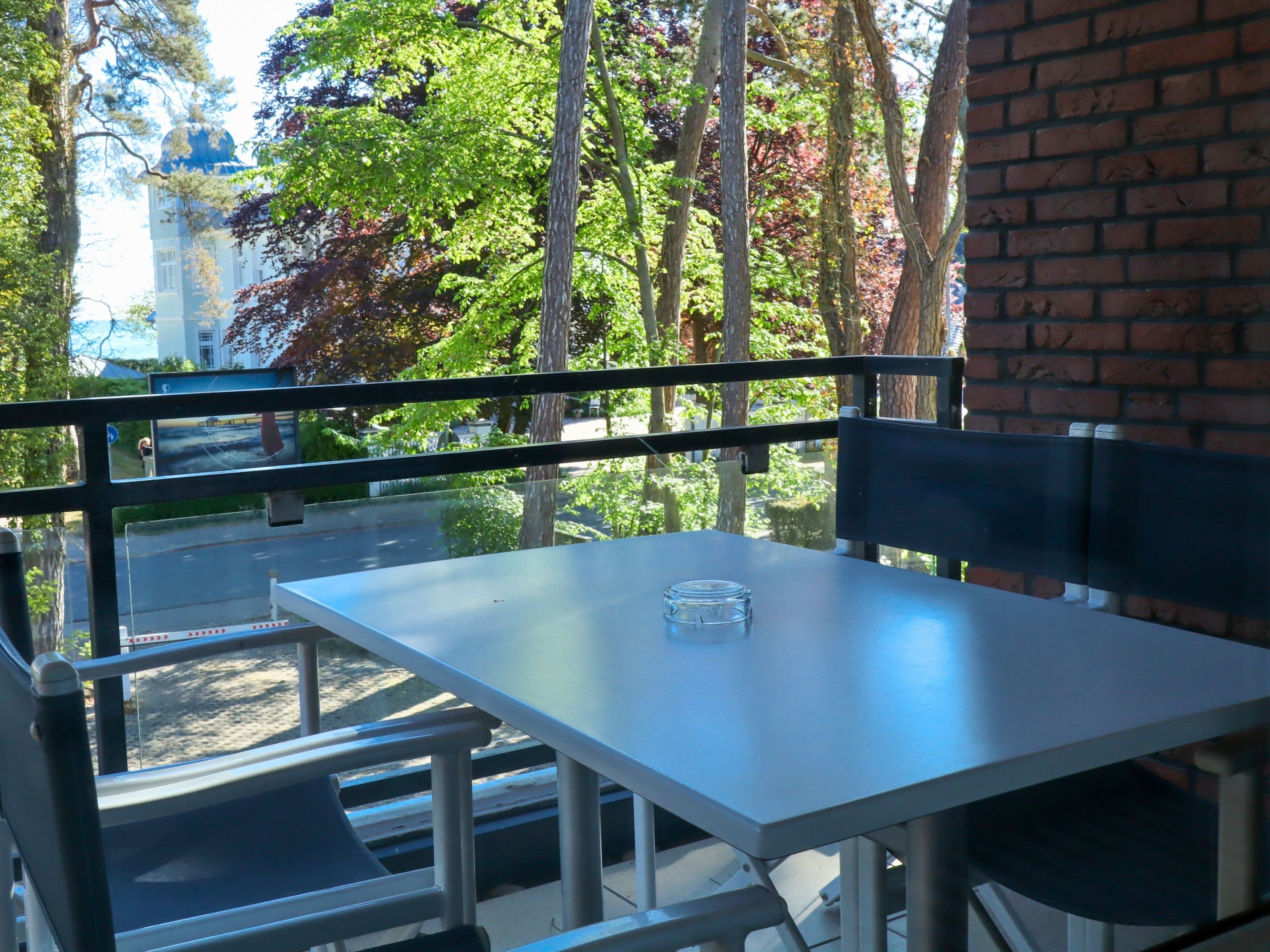 An outdoor seating area with a table and chairs, a glass of water, and a view of trees and a building in the background.