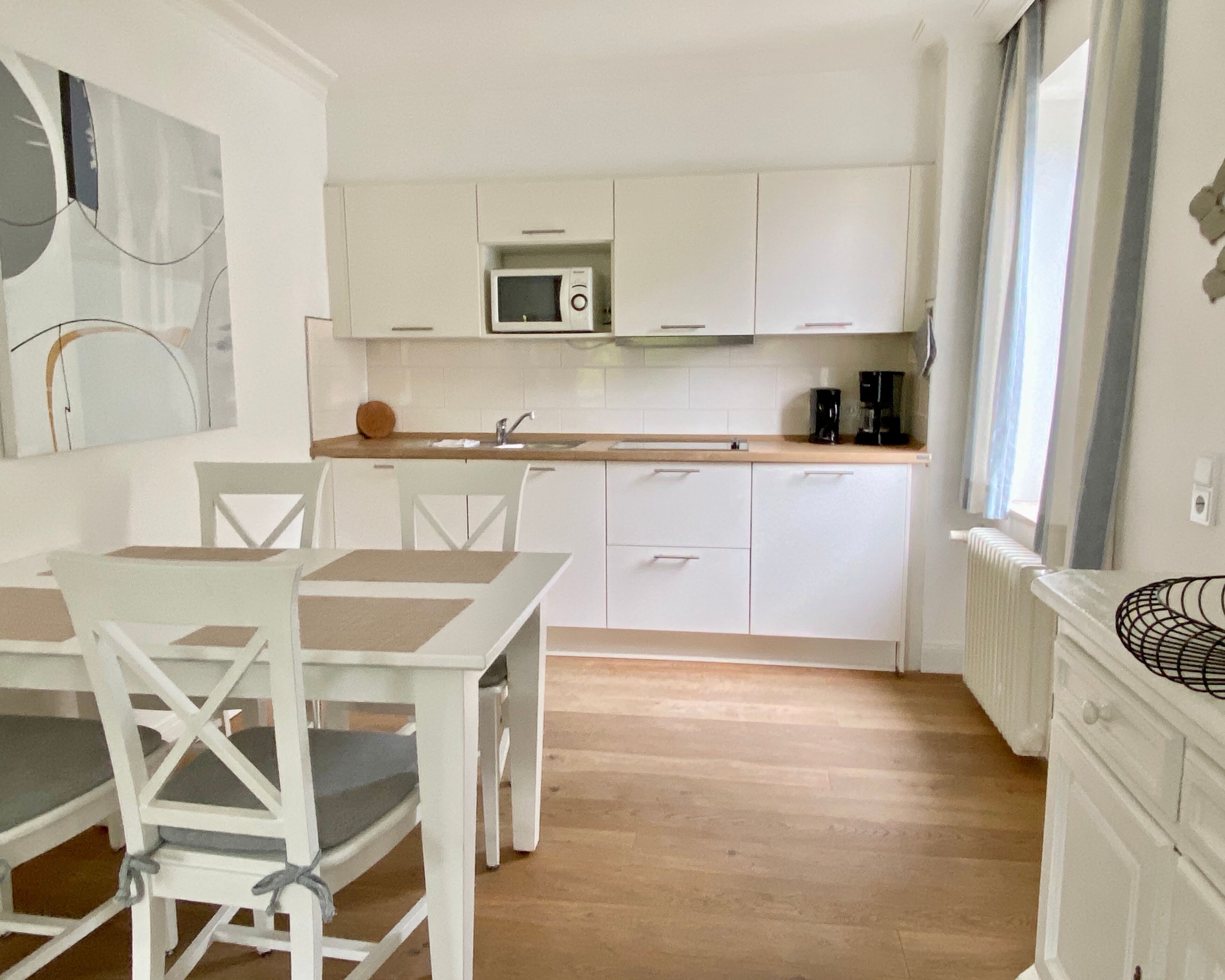 Une cuisine moderne avec des meubles blancs, un four à micro-ondes et une machine à café. On y trouve une table à manger avec des chaises et une fenêtre qui laisse entrer la lumière naturelle.