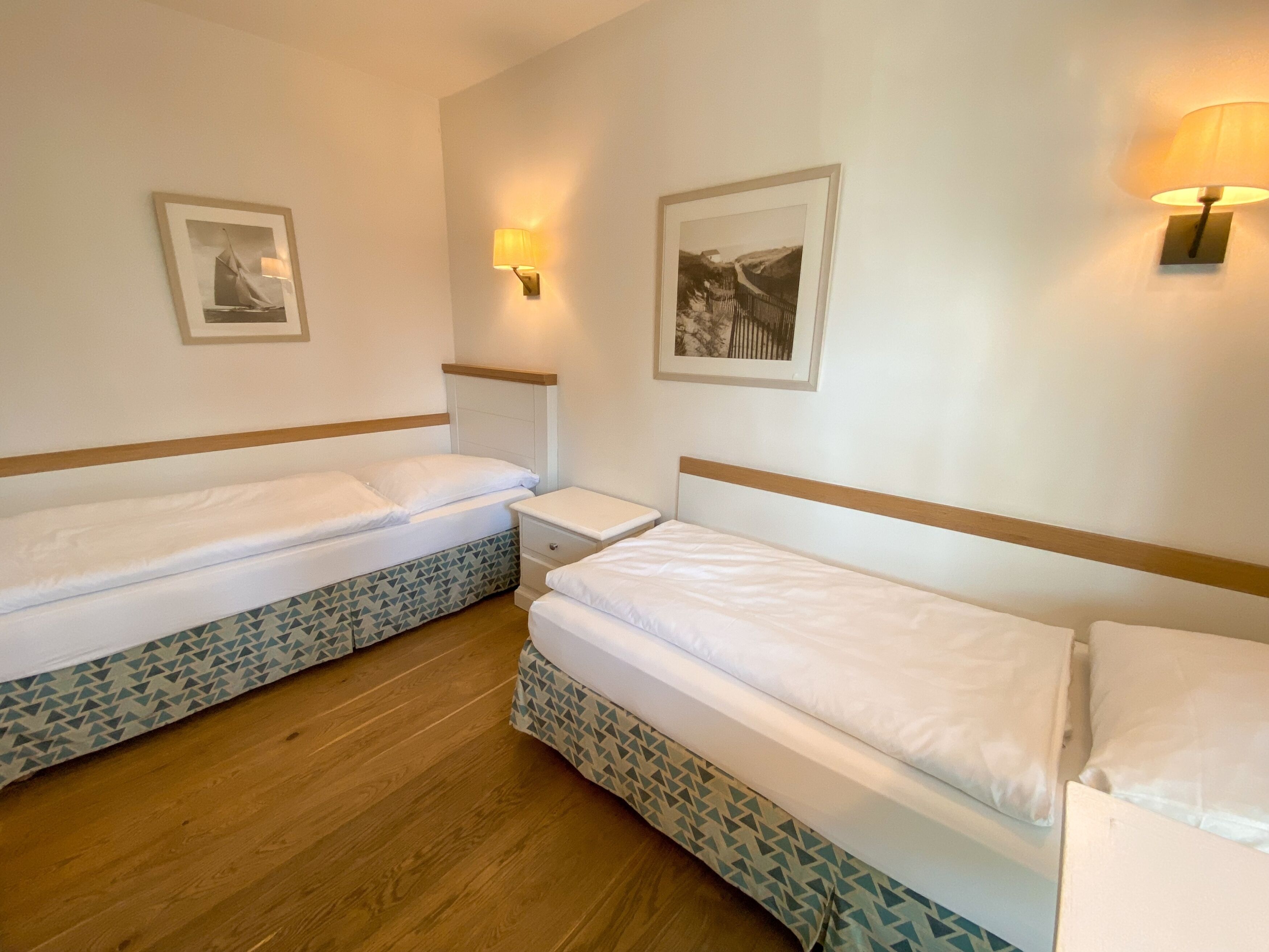 Une chambre d’hôtel avec deux lits simples, une table de chevet, une lampe fixée au mur et deux tableaux encadrés.