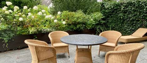 En uteplats med möbler i rotting och ett runt bord, omgiven av frodig grönska och vita blommor.