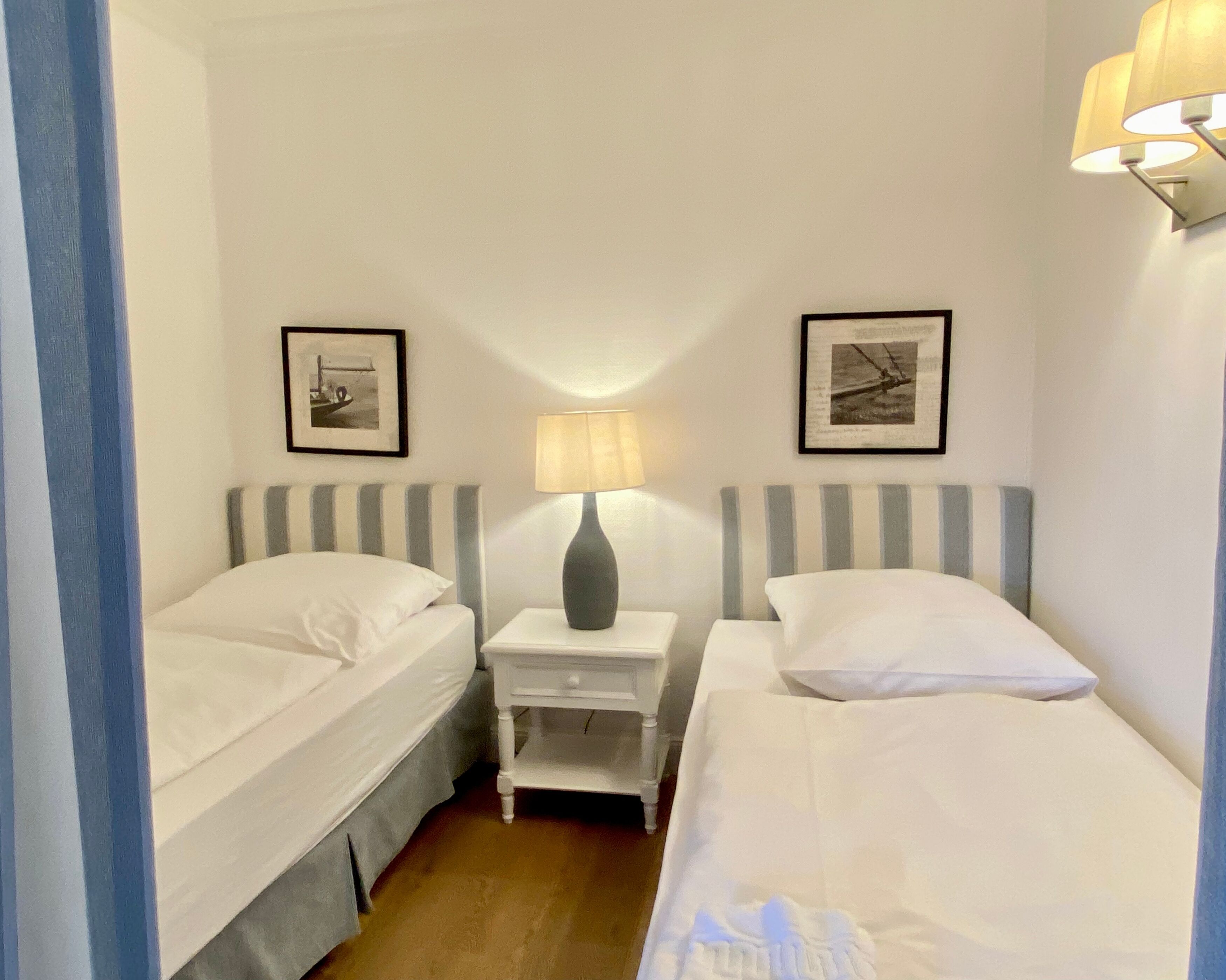 Habitación de hotel con dos camas individuales, una mesita de noche con lámpara y dos cuadros en las paredes.