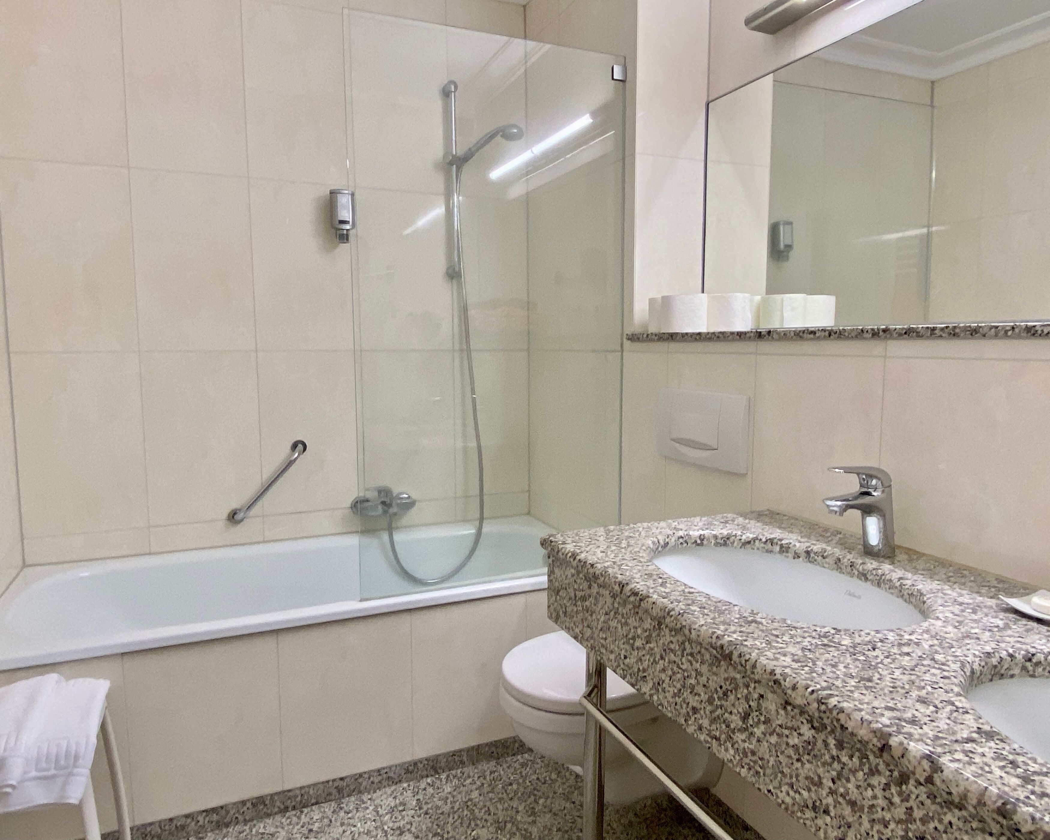Un baño con ducha envuelta en vidrio, inodoro blanco y lavamanos sobre encimera de granito.
