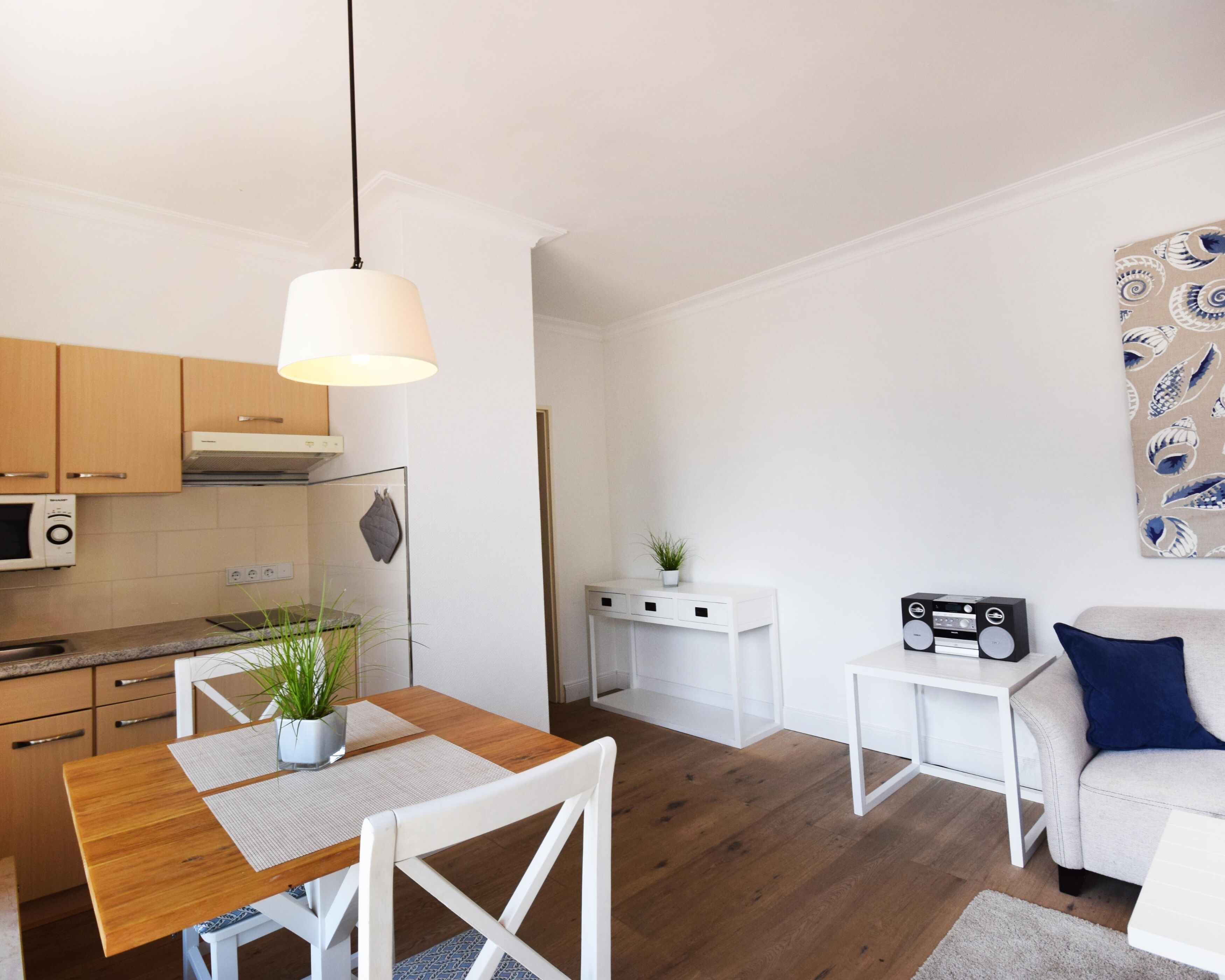 A modern living space with a wooden dining table, white chairs, a white sofa, a small white side table, and a kitchen area with wooden cabinets and a microwave.