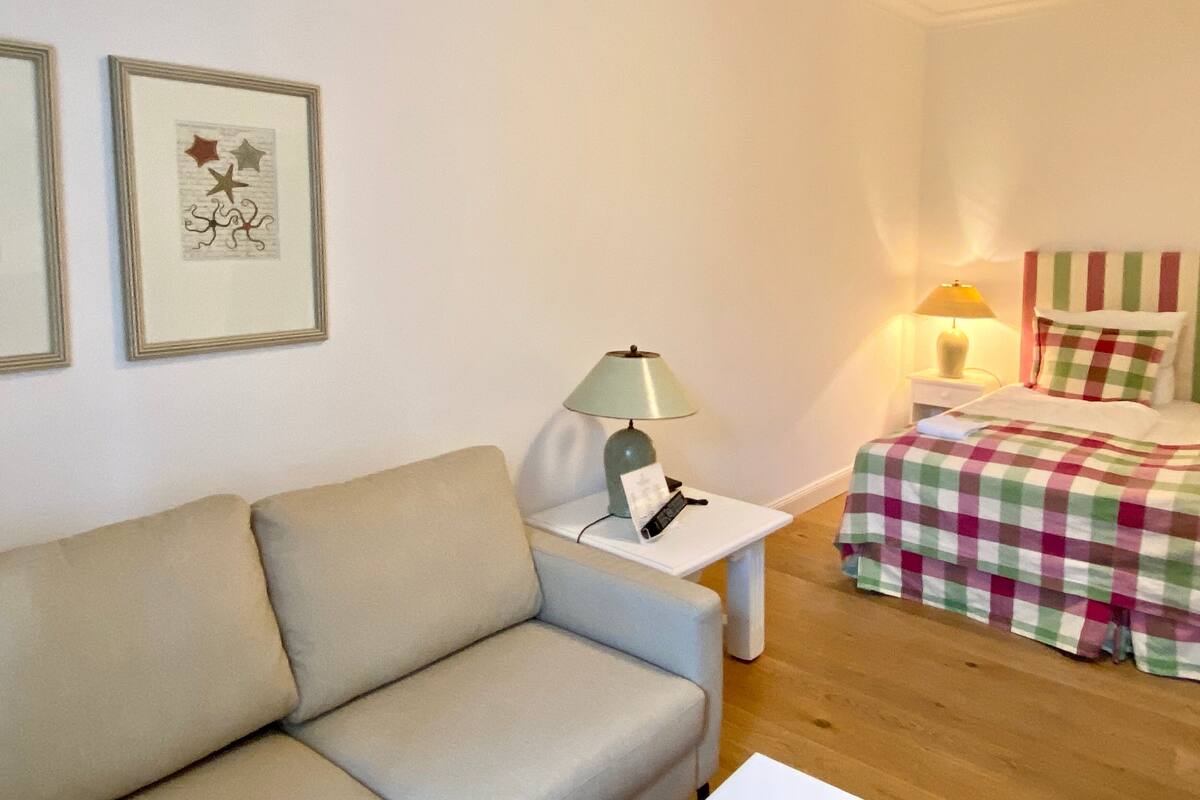 A room with a sofa, a bedside table with a lamp, a bed with a checkered blanket, and two framed pictures on the wall.