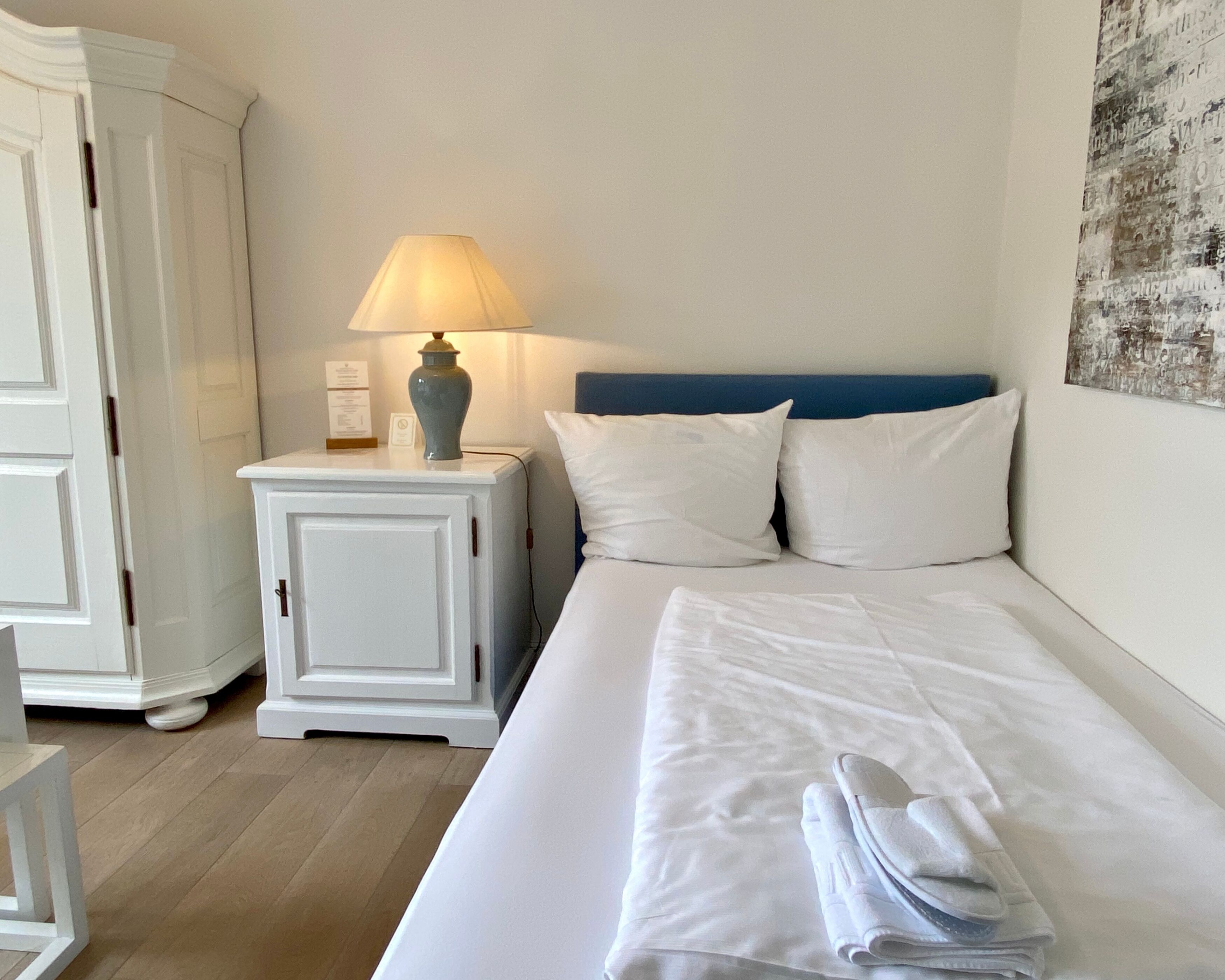 Una cama bien hecha con sábanas y almohadas blancas, una lámpara de noche y un pequeño mueble blanco con un jarrón encima.