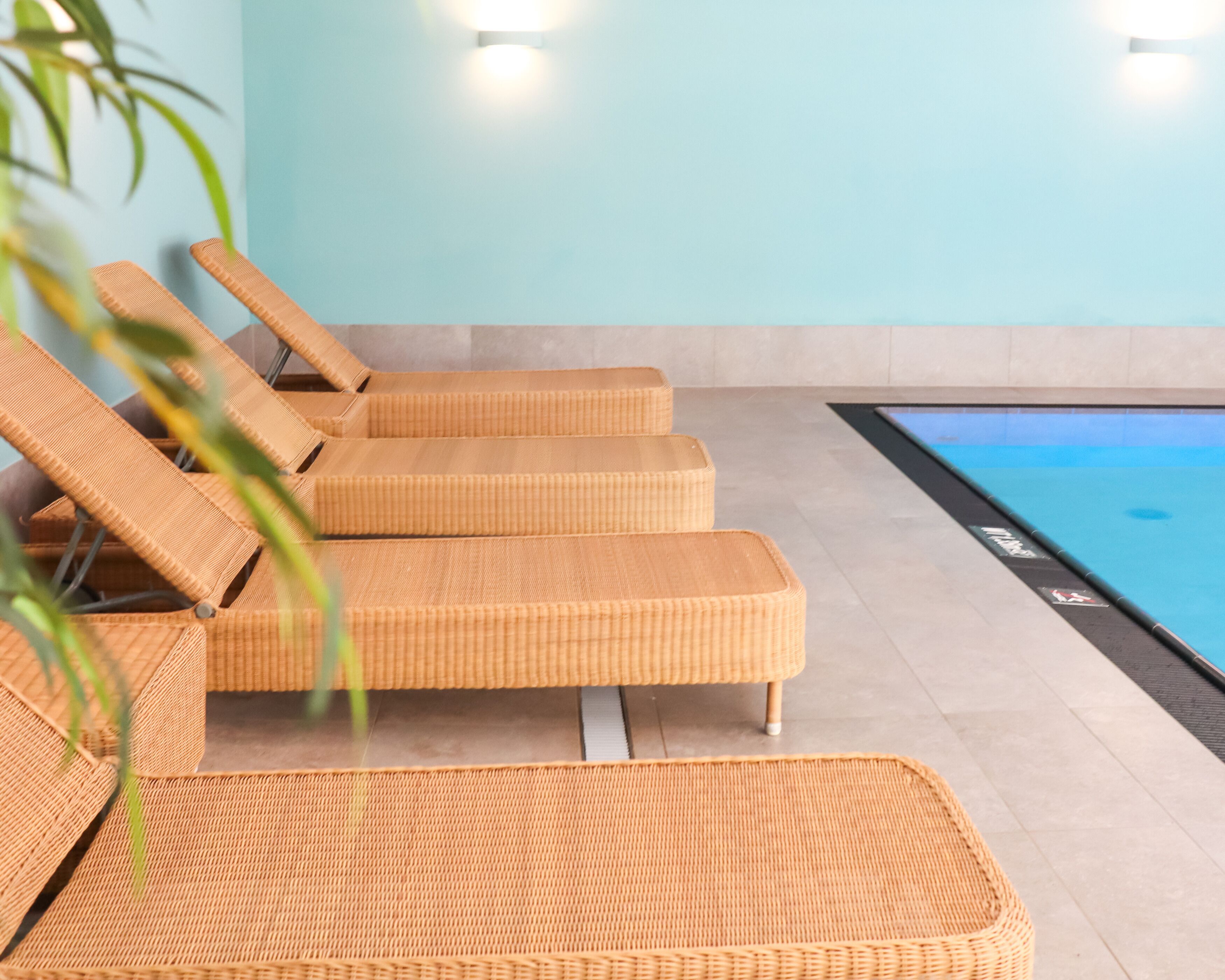 Une rangée de fauteuils de jardin en osier avec des armatures métalliques, une piscine et un mur bleu clair agrémenté de deux lampes murales.