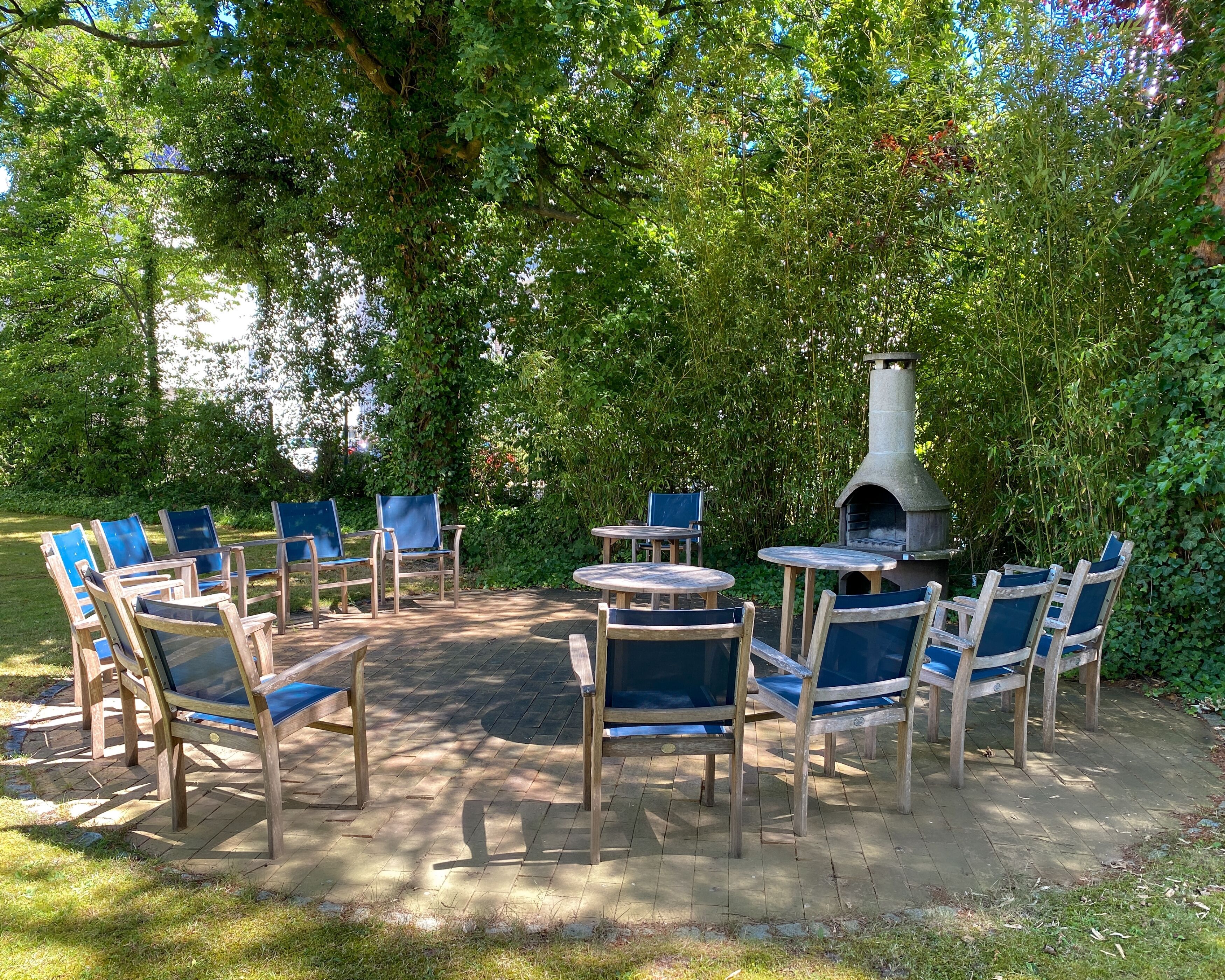 An outdoor seating area with wooden chairs and tables, surrounded by lush greenery and a stone-built outdoor oven.