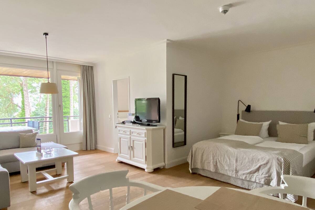 A hotel room with a bed, a television on a cabinet, a sofa, a table, and a view of trees through the window.