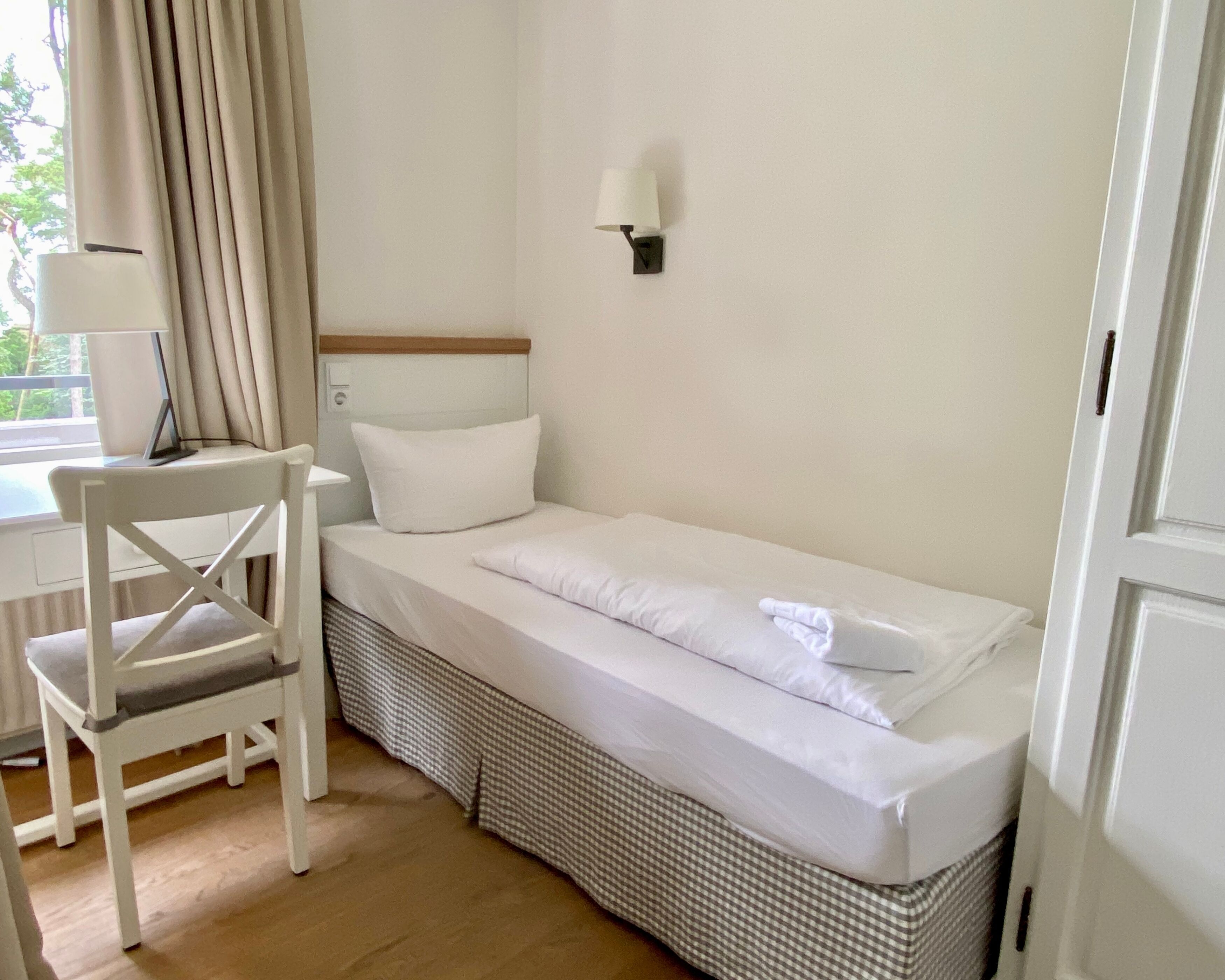 Ein Zimmer mit einem Einzelbett, einem karierten Bettdekor, einem weißen Stuhl, einem Schreibtisch mit Lampe und einer Wandleuchte.