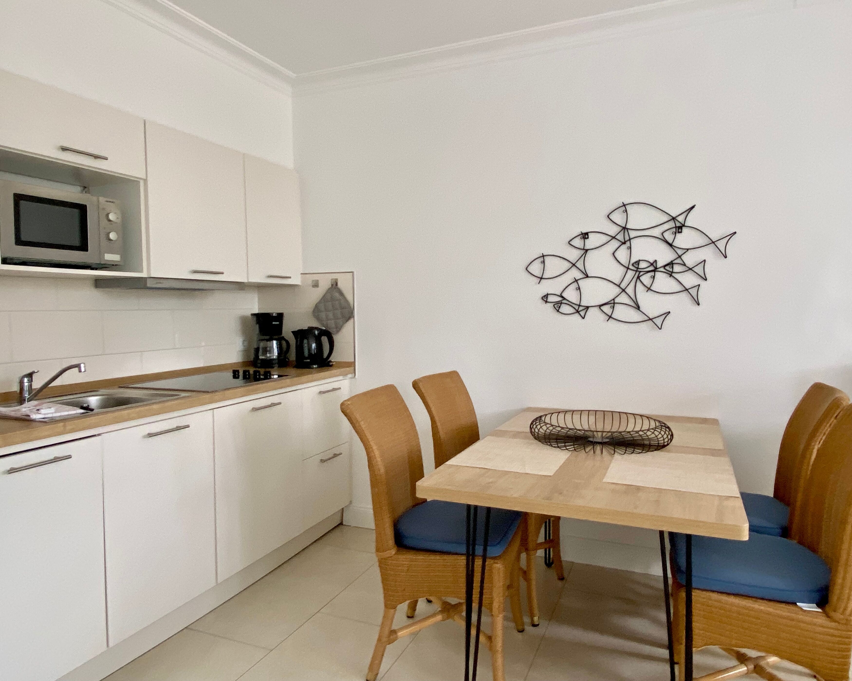 A compact kitchen with white cabinets, a microwave, and a coffee maker. There is a dining table with four chairs and a wall art of fish.