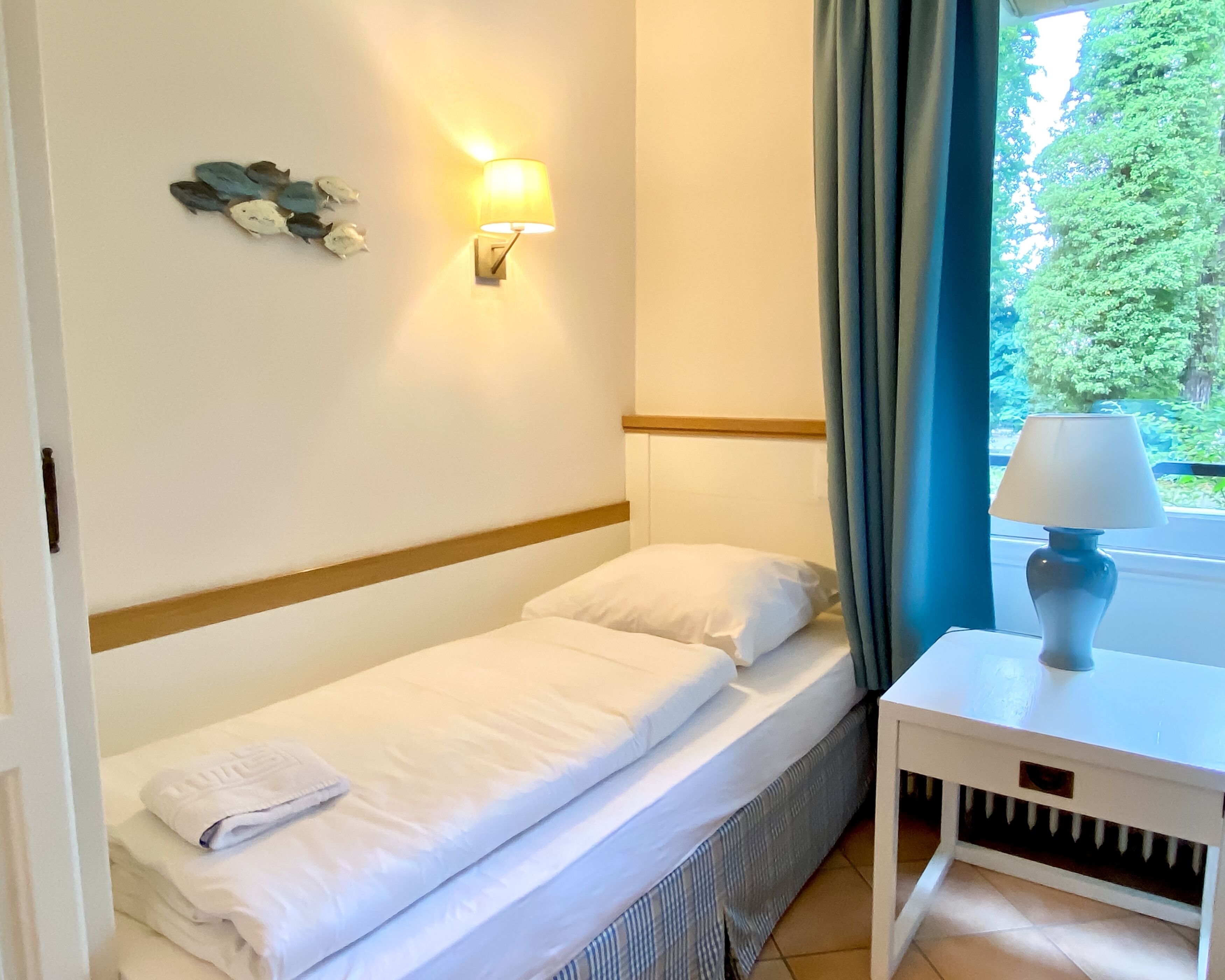 A single bed with white linens, a bedside table with a lamp, a wall-mounted light, and a view of greenery through a window with curtains.