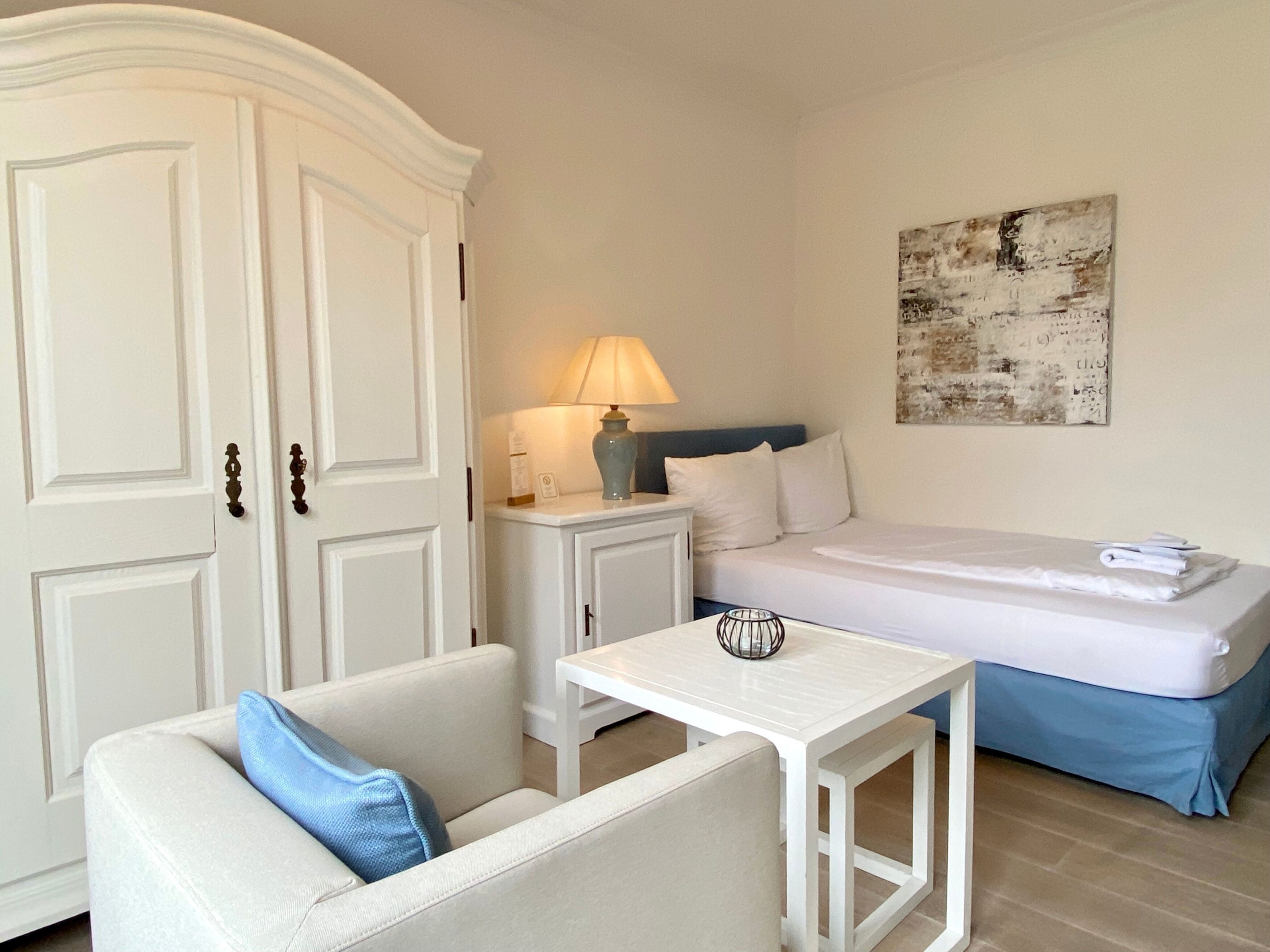 Un dormitorio con cama, una mesita de noche con lámpara, un armario blanco, una pequeña mesa y un cojín azul en un sofá.