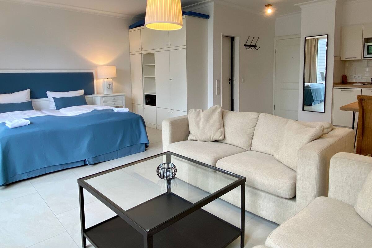 A modern hotel room with a bed, a sofa, a glass coffee table, and a kitchenette.
