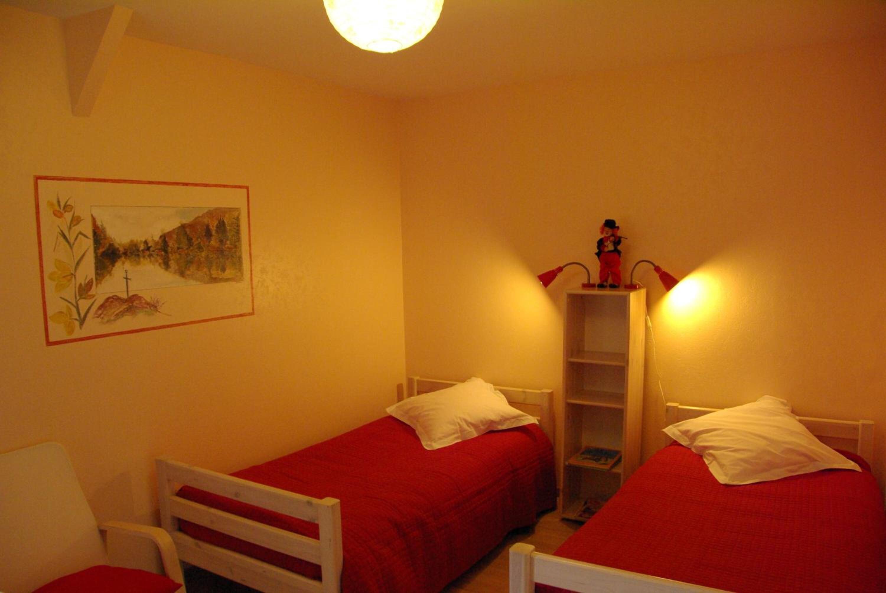 Una habitación con dos camas individuales, una silla blanca, una mesita con una lámpara roja y un estante de pared con una figurita.