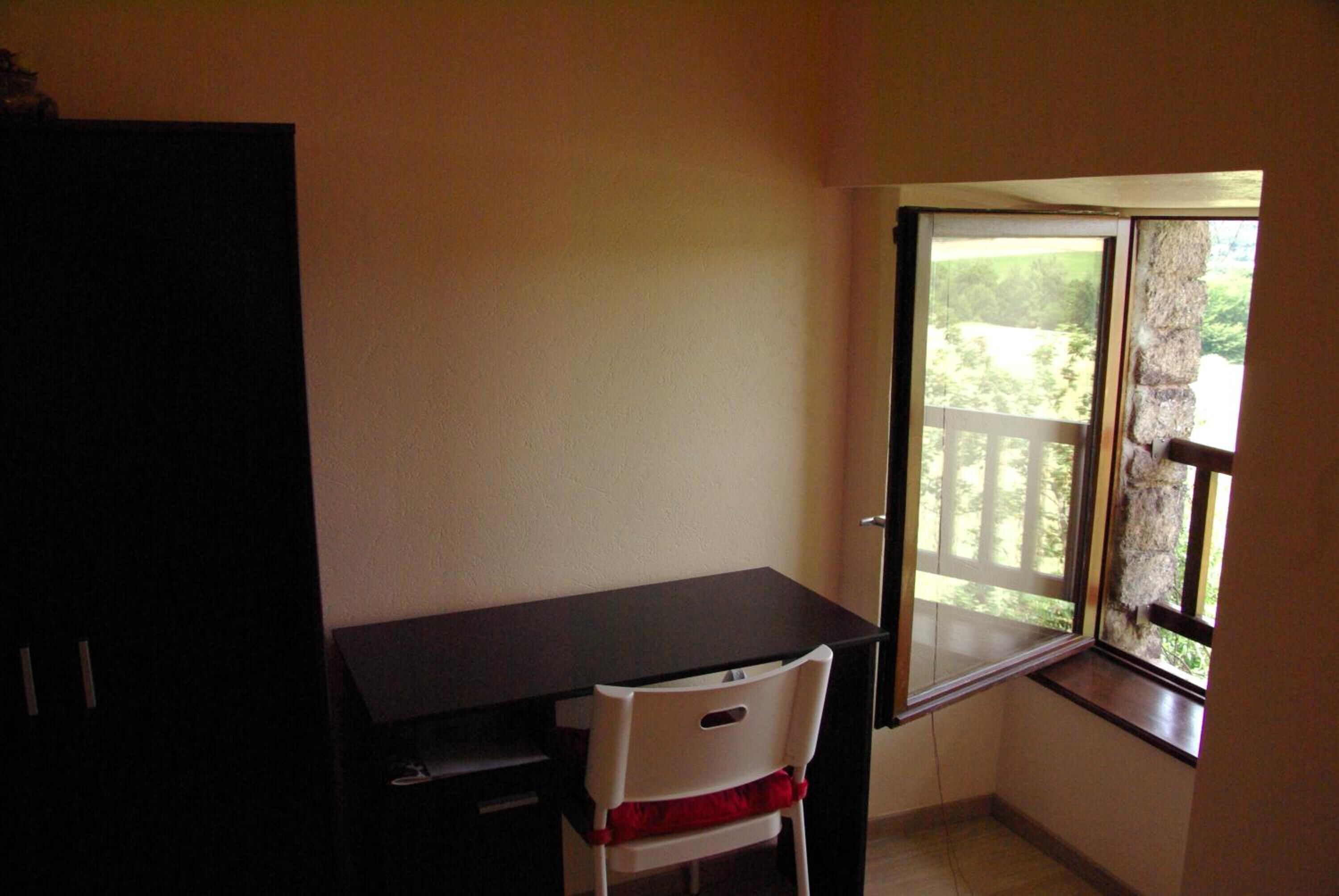 A small room with a desk and chair, a window with a view of greenery, and a door.