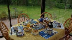 Een eettafel gedekt met borden, kommen en een theepot, omringd door rieten stoelen, met uitzicht op een tuin door grote ramen.