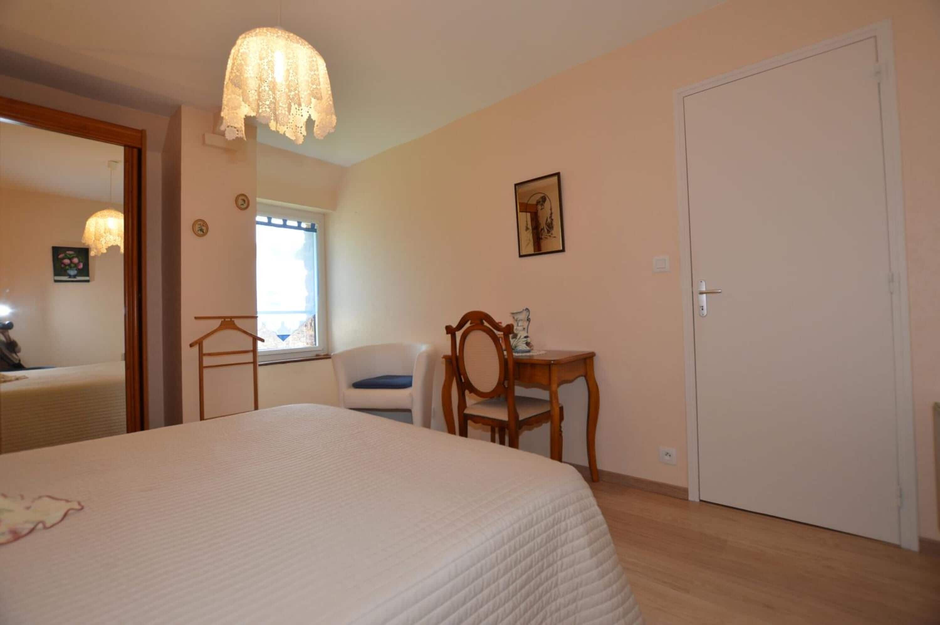 A bedroom with a large bed, a wooden chair, a small table, a hanging light fixture, a door, and a window.