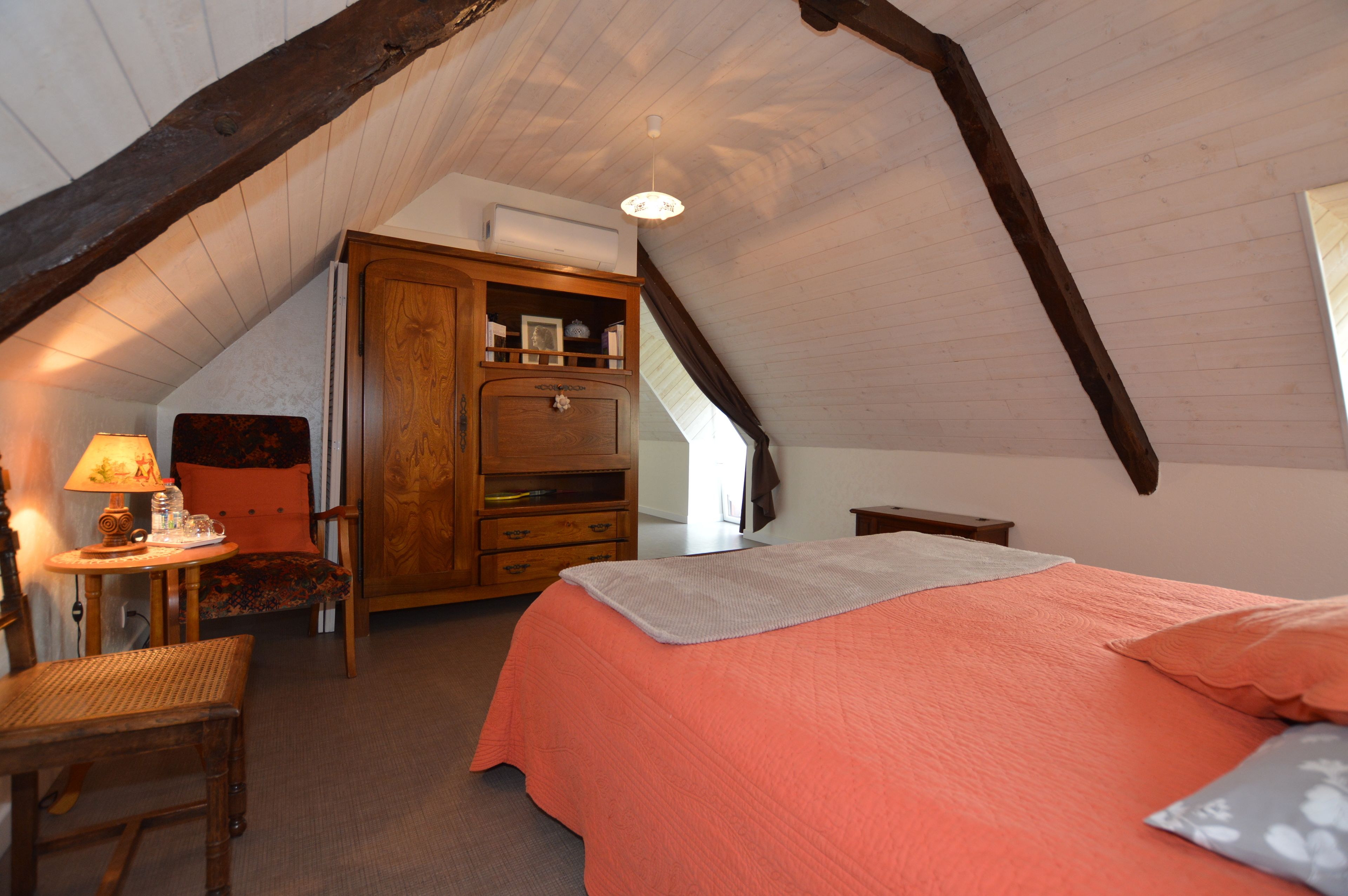 A cozy attic bedroom with a sloped ceiling, a bed with a pink bedspread, a wooden dresser, a chair, and a table with a lamp.