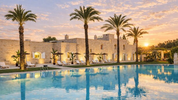 A pool with lounge chairs and palm trees, a stone building with arched windows, and a sunset in the background. - Masseria Antonio Augusto (Leverano)