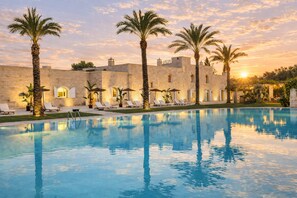 Una piscina con tumbonas y palmeras, un edificio de piedra con ventanas arqueadas y una puesta de sol al fondo.