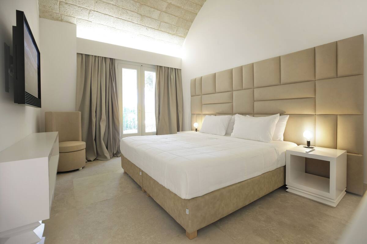 Een moderne hotelkamer met een groot bed, een nachtkastje met een lamp, een aan de muur bevestigde televisie en een raam met gordijnen.