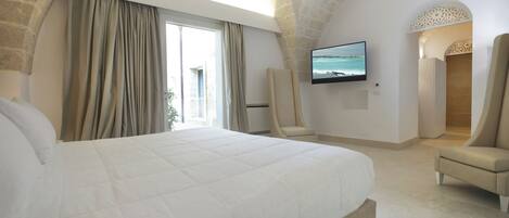 A spacious bedroom with a large bed, a flat-screen TV mounted on the wall, and a view of the sea through the window.