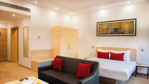 Une chambre d’hôtel moderne comprenant un lit, un canapé, une table de chevet, une petite table, un tableau et un panier de fruits posé sur une table basse.