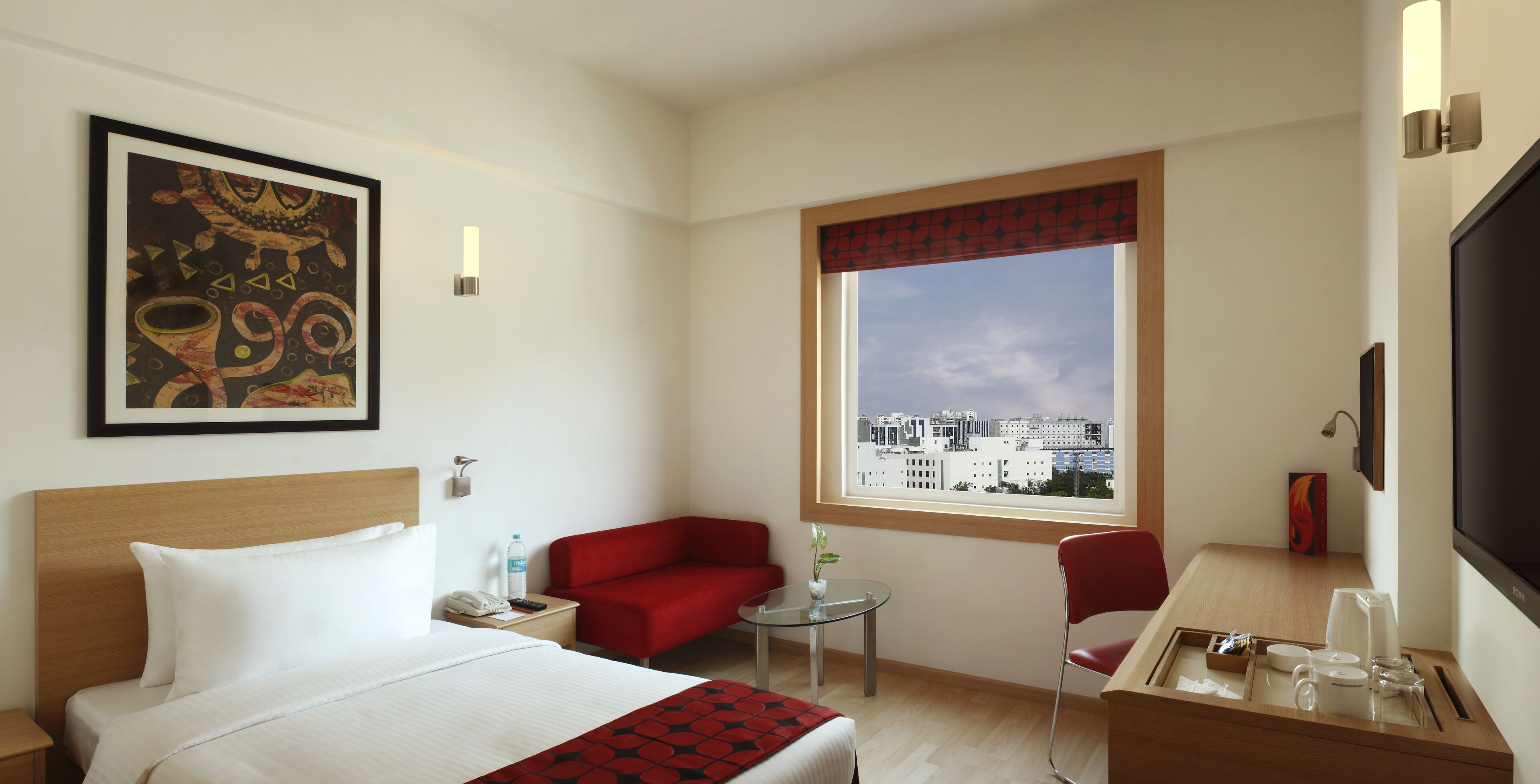 Una habitación de hotel con una cama, un sillón rojo, una mesita con un jarrón, una ventana con vistas a la ciudad y un televisor en la pared.