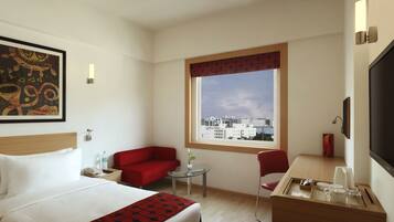 Una habitación de hotel con una cama, un sillón rojo, una mesita con un jarrón, una ventana con vistas a la ciudad y un televisor en la pared.