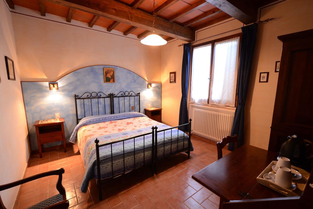 Ein Schlafzimmer mit Bett, Nachttischen, einem Holzstuhl, einem Esstisch mit Tassen und einem Wasserkocher, sowie einem Fenster mit Vorhängen.