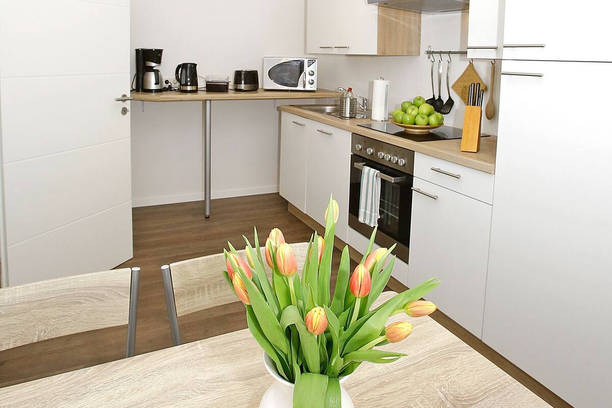 Une cuisine moderne avec des armoires blanches, un four encastré, un four à micro-ondes et une machine à café. Un vase de tulipes est posé sur la table à manger.