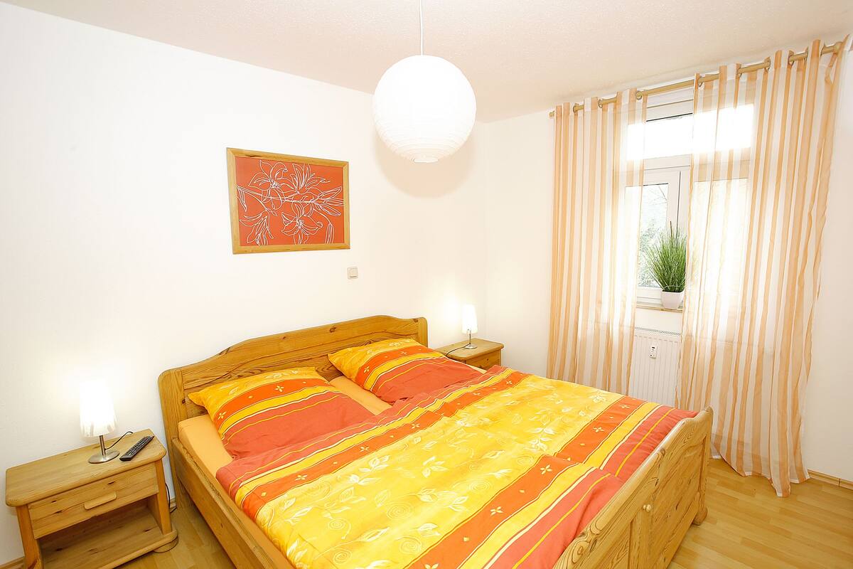 Un lit bien fait, recouvert d’une couvre-lit à rayures, une table de chevet avec une lampe, une œuvre d’art accrochée au mur et une fenêtre avec des rideaux.