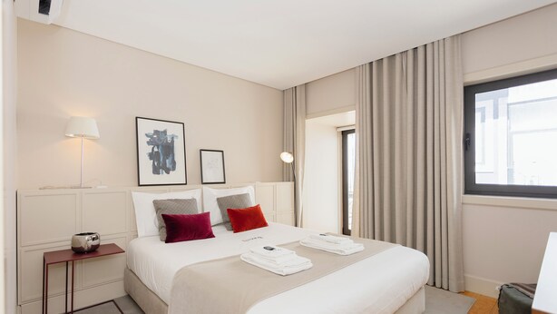 Quarto de hotel com cama, mesa de cabeceira, luminária e janela com cortinas.