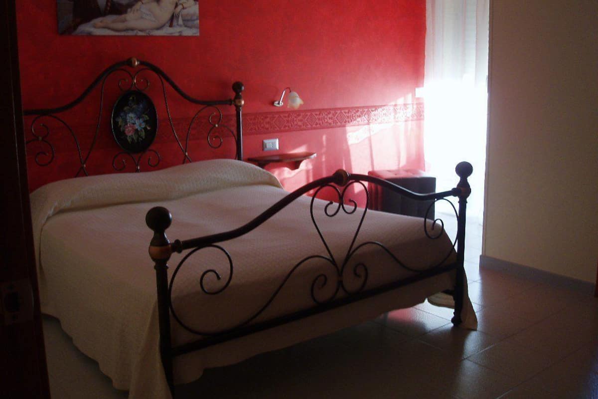 Ein Bett mit Metallgestell und weißer Bettdecke in einem Zimmer mit roten Wänden und einem Gemälde darüber.