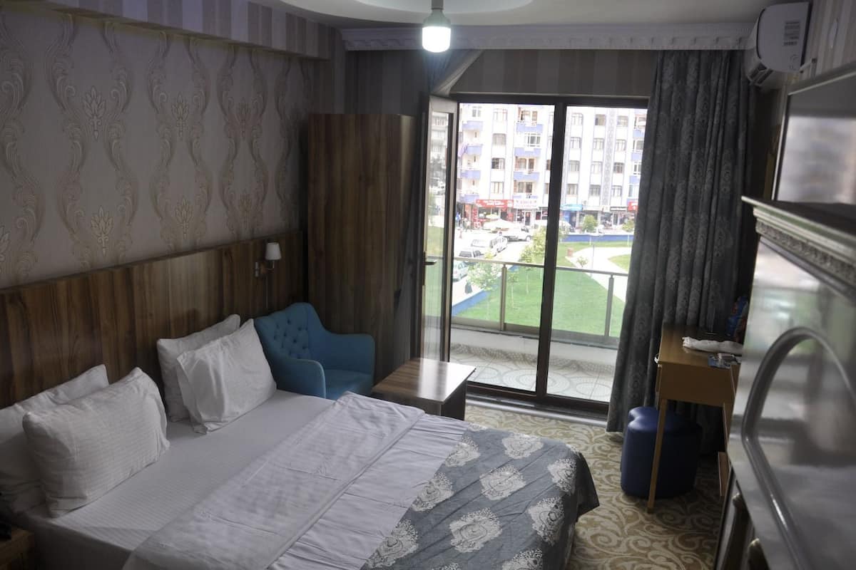 A hotel room with a large bed, a blue armchair, a small table, and a view of a building and greenery outside.