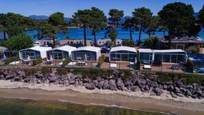 Una zona costera con cabañas modernas de techo de cristal y con asientos exteriores, un litoral rocoso y vistas al mar y montañas al fondo.
